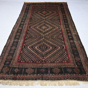May include: A rectangular rug featuring a central diamond pattern in brown and black, framed by a repeating geometric border. The rug has a red accent and fringed edges, displaying a traditional design.