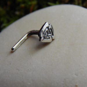 Pode incluir: Um piercing de nariz prateado com um design de nó celta. O piercing é dobrado em ângulo reto e tem um pequeno nó decorativo na extremidade. O piercing está sobre uma pedra branca lisa.