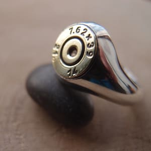 Puede incluir: Un anillo de plata con una vaina de bala de color dorado en la parte superior. La vaina de bala tiene los números "7.62x39" grabados en ella.