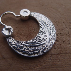 Könnte beinhalten: Silberner Creolenohrring mit einem filigranen Design aus Blättern und einem Kreuz. Der Ohrring ist halbmondförmig und hat eine polierte Oberfläche.