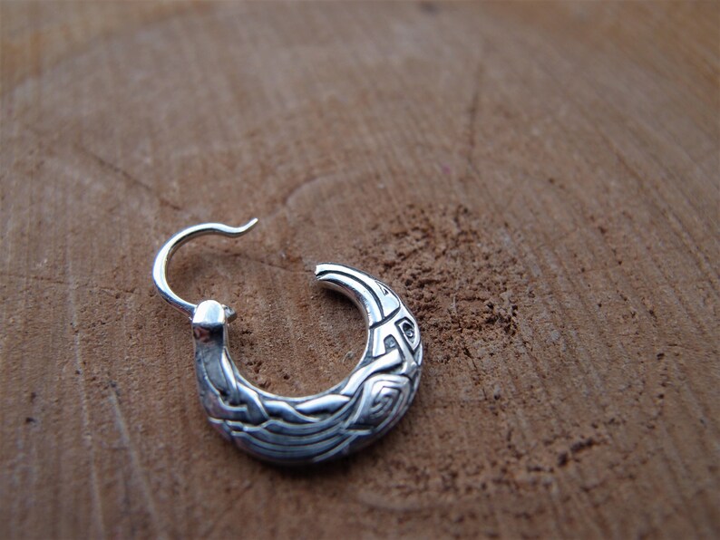 May include: Silver hoop earring with a detailed design of a bird or animal with intricate patterns. The earring is on a wooden surface.