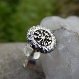 Puede incluir: Un pendiente de plata con un borde texturizado y un centro negro con un diseño de brújula vikinga blanca. El pendiente se encuentra sobre un fondo borroso verde y blanco.