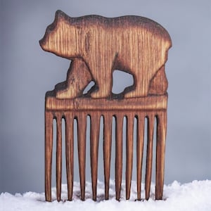 May include: A wooden comb with a carved bear design on the top. The comb is brown and has a natural wood finish. It is sitting on a white surface.