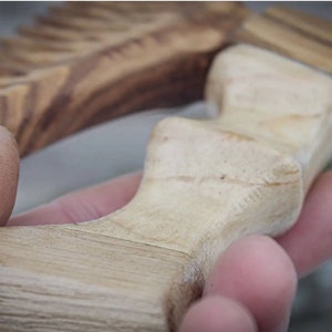 May include: Close-up of a wooden handle with a light brown color and a smooth, polished finish. The handle has a unique, curved shape with a series of ridges.