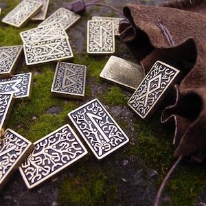 May include: Gold-coloured rune stones with etched symbols are scattered on green moss. A brown leather pouch is in the background. The rectangular stones feature detailed patterns and symbols, suggesting their use in divination or spiritual practices.