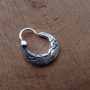 May include: A silver hoop earring with a detailed, intricate design. The earring is made of metal and has a smooth, polished finish.