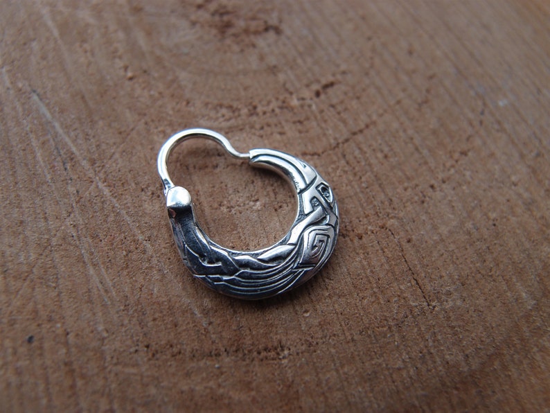 May include: A silver hoop earring with a detailed, intricate design. The earring is made of metal and has a smooth, polished finish.