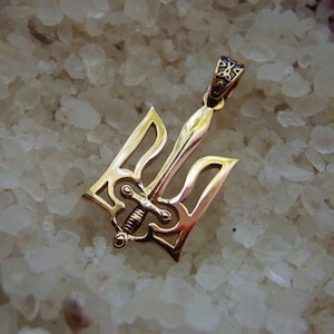 Może przedstawiać: Złoty wisiorek w kształcie ukraińskiego trójzębu, tradycyjnego symbolu Ukrainy. Wisiorek jest ozdobiony misternymi rzeźbami i małym mieczem w środku.