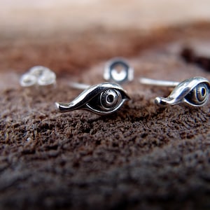 Puede incluir: Un par de pendientes de plata con forma de ojo. Cada pendiente presenta un diseño de ojo detallado con una pupila en relieve.