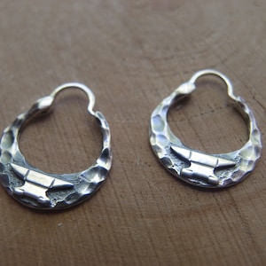 May include: A pair of silver hoop earrings with a hammered texture and an anvil design.