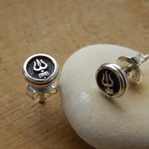 May include: Silver stud earrings with a black trident symbol. The earrings are round with a raised border and a post for piercing. One earring is on a white stone, and the other is on a wooden surface.