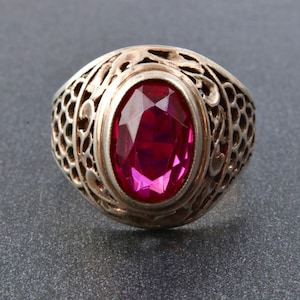May include: A silver ring featuring an oval, faceted, ruby-red gemstone. The ring has an open filigree design around the gemstone, with a detailed, decorative pattern. The ring is set against a grey background.