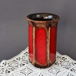 Puede incluir: Un jarrón alto y cilíndrico en rojo y marrón. El jarrón presenta paneles verticales de color rojo brillante, enmarcados por bandas marrones. El borde y el interior son de color marrón oscuro. El jarrón es probablemente de cerámica y está sobre un mantel de encaje blanco. Altura: unos 20 cm.