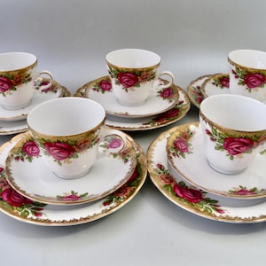 Könnte beinhalten: Ein sechsteiliges Teeservice mit weißen Tassen und Untertassen. Jedes Teil hat einen goldenen Rand und ein Rosenmuster in Rot und Rosa. Das Set wird auf einer hellgrauen Oberfläche präsentiert.