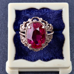 May include: A silver-toned ring featuring a large, oval-cut, deep red gemstone set in a decorative, filigree-style metal band. The ring is displayed inside a square, cream-colored box with a plush, dark blue velvet insert.