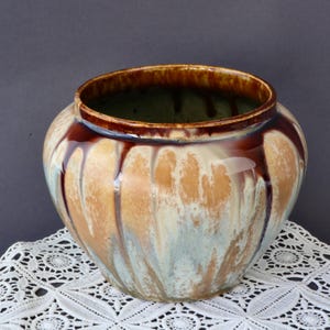 Peut inclure: Un vase en céramique avec un corps large et arrondi et un bord marron foncé. L'extérieur présente un mélange de glaçure beige, bleu clair et marron, avec un effet dégoulinant. Le vase repose sur une nappe en dentelle blanche.