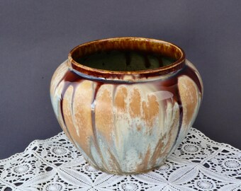 Pode incluir: Um vaso de cerâmica com um corpo largo e arredondado e uma borda marrom escura. O exterior apresenta uma mistura de esmalte bege, azul claro e marrom, com um efeito de gotejamento. O vaso está sobre uma toalha de renda branca.