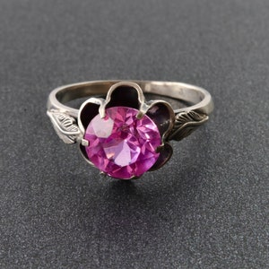 Op de afbeelding: Een zilveren ring met een roze edelsteen in een bloemvormig ontwerp.