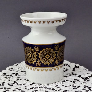 Op de afbeelding: Een witte keramische vaas met een uitlopende boven- en onderkant, met een donkerblauwe band met gouden cirkelvormige en bloemmotieven. Gouden randen accentueren de boven- en onderkant. De vaas wordt gepresenteerd op een wit kanten kleedje.