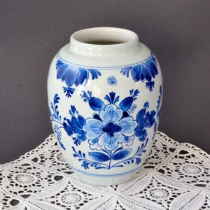 Può includere: Un vaso di ceramica bianca con motivi floreali blu. Il vaso ha un corpo arrotondato e un collo stretto. Il design floreale blu include grandi fiori e piccoli accenti di foglie. Il vaso è esposto su una tovaglietta di pizzo bianco.