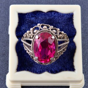 Può includere: Un anello color argento con una grande pietra preziosa rosa vibrante dal taglio ovale, incastonata in una fascia metallica decorativa in stile filigrana. L'anello è appoggiato su un cuscinetto di velluto blu scuro in un portagioie quadrato color crema.