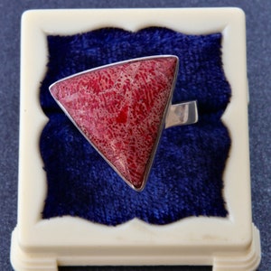 Pode incluir: Um anel com uma grande pedra vermelha de formato triangular com um padrão texturizado de veios brancos, engastada numa banda de metal de tom prateado polido. A pedra apresenta detalhes de superfície naturais e intrincados.
