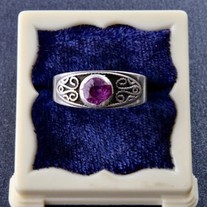 Può includere: Un anello in argento con una pietra preziosa viola rotonda e sfaccettata al centro. La fascia presenta intricati motivi a volute metalliche simmetriche su entrambi i lati della pietra, creando un design dettagliato sulla parte anteriore.