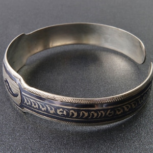 Op de afbeelding: Zilverkleurige manchetarmband met een donkerblauwe emaille band met een sierlijk patroon. De open armband wordt gepresenteerd op een donkergrijze ondergrond. De manchet is ongeveer 5 cm breed.