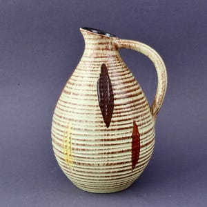 Peut inclure: Une carafe en céramique avec une base de couleur crème et des lignes horizontales brunes en relief. Le récipient présente un corps arrondi, une anse incurvée et un bord intérieur sombre. Des motifs verticaux en forme de feuille sont répartis.