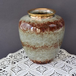 May include: A decorative vase with a wide body and a gold rim. The vase has a textured, light green and brown pattern. The vase is sitting on a white lace tablecloth. The vase is a decorative home decor item.