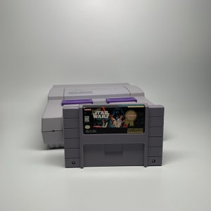 May include: A Super Nintendo Entertainment System console with a Star Wars video game cartridge. The game cartridge features a colorful image of characters from the movie. The console is light gray with purple accents.