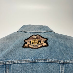 May include: A blue denim jacket with a Harley Owners Group (H.O.G.) patch on the back. The patch features an eagle with wings spread and a motorcycle wheel in the center.