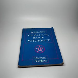 May include: A blue paperback book titled Buckland's Complete Book of Witchcraft by Raymond Buckland. The cover features a white pentagram inside a pink circle, with white text and thin pink rectangular borders on a solid blue background.