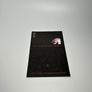 May include: A black and white comic book cover with the title "THE BLAIR WITCH PROJECT."  The cover features a close-up of a person's face wearing a white hat, with only the eyes visible. The background is black with a red symbol of a stick figure.