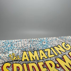Puede incluir: Una portada de c&oacute;mic con el t&iacute;tulo The Amazing Spider-Man en letras amarillas en negrita con un contorno rojo. El fondo muestra un patr&oacute;n detallado de telara&ntilde;a azul sobre una superficie blanca, con una peque&ntilde;a mancha marr&oacute;n en el borde superior.