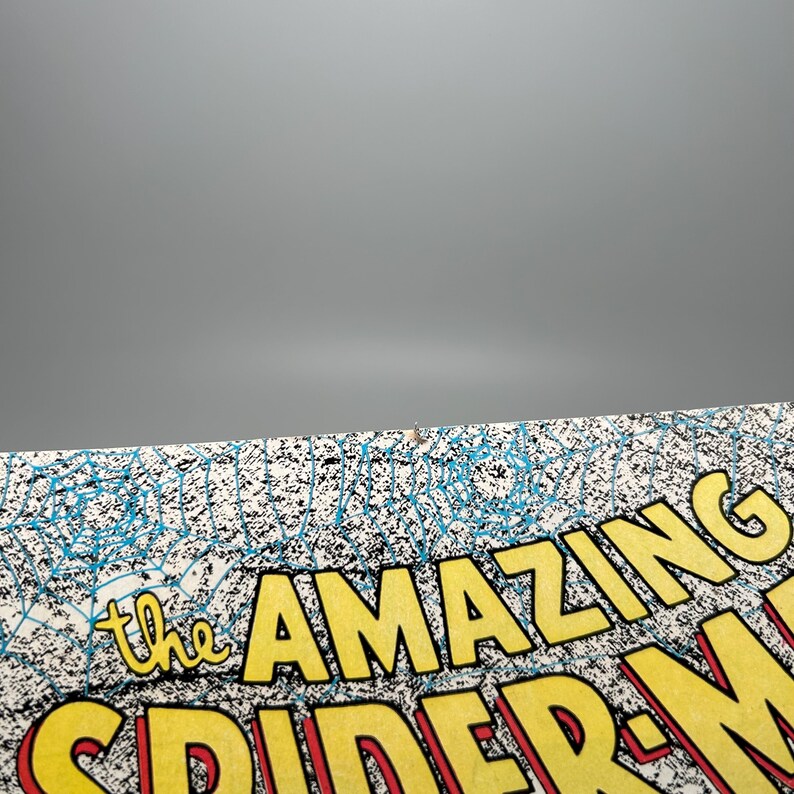 Puede incluir: Una portada de c&oacute;mic con el t&iacute;tulo The Amazing Spider-Man en letras amarillas en negrita con un contorno rojo. El fondo muestra un patr&oacute;n detallado de telara&ntilde;a azul sobre una superficie blanca, con una peque&ntilde;a mancha marr&oacute;n en el borde superior.