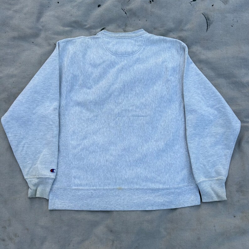 May include: A light grey Champion sweatshirt with a crew neck and long sleeves. The Champion logo is visible on the left sleeve cuff. The sweatshirt has a ribbed collar, cuffs, and hem.