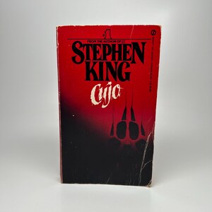 May include: A vintage paperback book titled "Cujo" by Stephen King. The cover is predominantly red with black text and a graphic of claw marks. The book is from the author of "It".