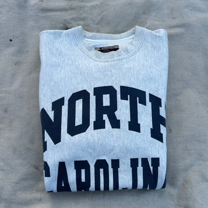 May include: A light grey Champion sweatshirt with the words "NORTH CAROLINA" printed in navy blue block letters. The sweatshirt is folded and appears to be made of a soft, comfortable material.