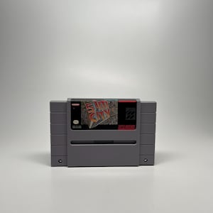 May include: A gray Super Nintendo video game cartridge for SimCity. The label features the game's title in a stylized font, with a cityscape graphic. The cartridge is in good condition, with a clean, minimalist design.