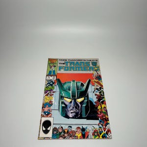 May include: A vintage The Transformers comic book issue number 22, featuring a large green robot head illustration on the cover. The title reads More Than Meets The Eye The Transformers. The cover art includes various colorful character figures.