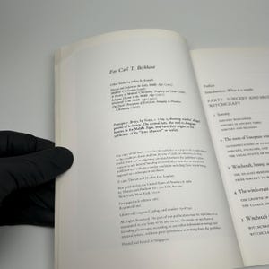 Op de afbeelding: Open boekpagina's met tekst over hekserij en tovenarij. Het boek heet "Sorcery and Witchcraft" en bevat secties over tovenarij wereldwijd en in de oudheid. Het boek wordt vastgehouden door een gehandschoende hand.