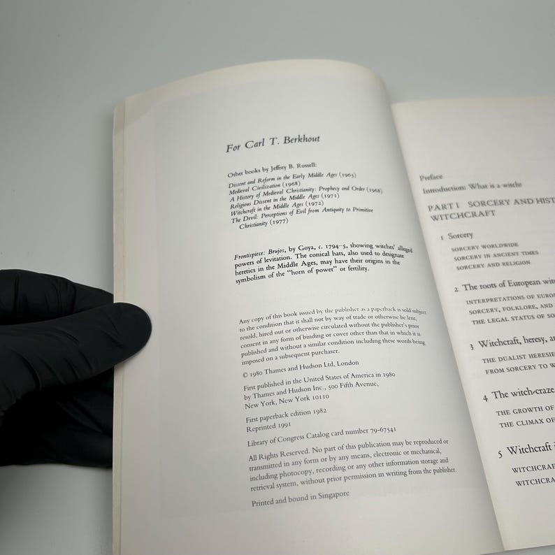 Op de afbeelding: Open boekpagina's met tekst over hekserij en tovenarij. Het boek heet "Sorcery and Witchcraft" en bevat secties over tovenarij wereldwijd en in de oudheid. Het boek wordt vastgehouden door een gehandschoende hand.