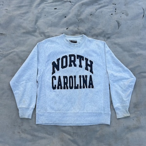 May include: A light gray crewneck sweatshirt with the words "NORTH CAROLINA" in navy blue arched across the chest. The sweatshirt has long sleeves and a ribbed collar and cuffs. The fabric appears to be a soft, comfortable material.