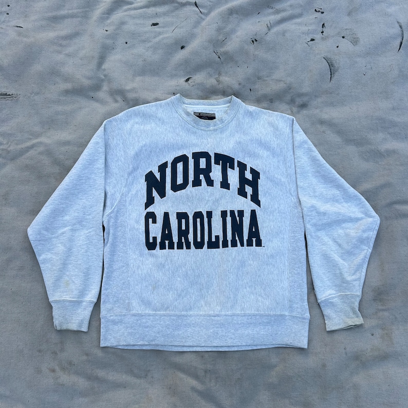 May include: A light gray crewneck sweatshirt with the words "NORTH CAROLINA" in navy blue arched across the chest. The sweatshirt has long sleeves and a ribbed collar and cuffs. The fabric appears to be a soft, comfortable material.
