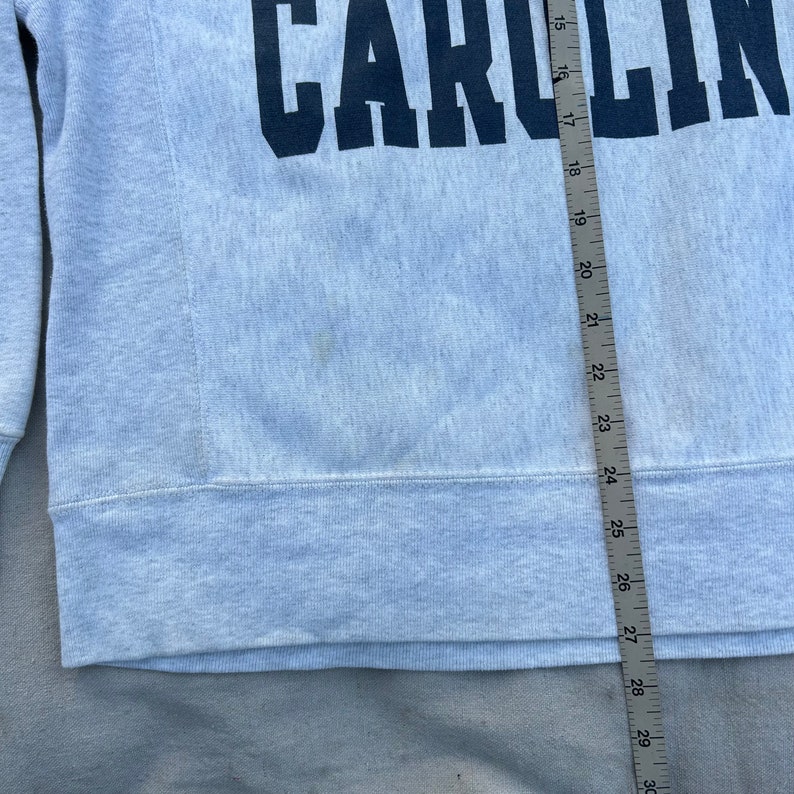 May include: Light gray sweatshirt with the word "CAROLIN" printed in navy blue across the chest. The sweatshirt is measured with a ruler, showing a length of approximately 15 to 30 inches. The fabric appears to be a soft, textured material.
