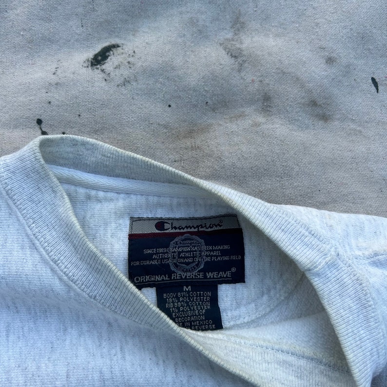 May include: Close-up of a light gray Champion sweatshirt. The tag reads "ORIGINAL REVERSE WEAVE" and includes the brand logo. The sweatshirt is a medium size and made of cotton and polyester.
