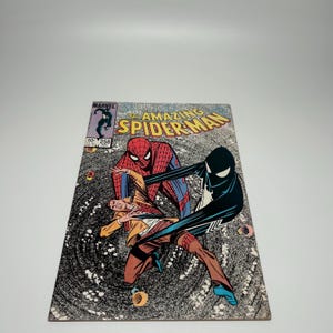 Puede incluir: El c&oacute;mic The Amazing Spider-Man n&uacute;mero 258, con una ilustraci&oacute;n de portada de Spider-Man con traje rojo y azul y una figura con traje negro interactuando con una persona con chaqueta beige y pantalones marrones sobre un fondo negro arremolinado.