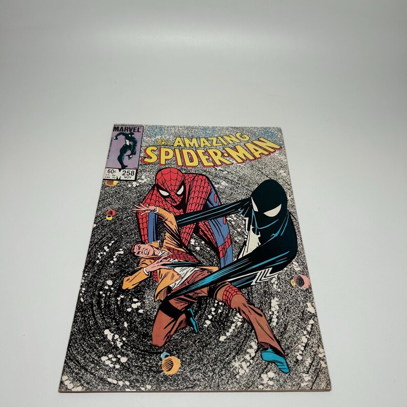 Puede incluir: El c&oacute;mic The Amazing Spider-Man n&uacute;mero 258, con una ilustraci&oacute;n de portada de Spider-Man con traje rojo y azul y una figura con traje negro interactuando con una persona con chaqueta beige y pantalones marrones sobre un fondo negro arremolinado.