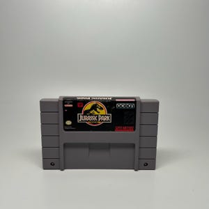 May include: A gray Super Nintendo video game cartridge for Jurassic Park. The label features a black and yellow circular logo with a dinosaur silhouette, the text Jurassic Park, the Ocean logo, and the Super Nintendo Entertainment System branding.